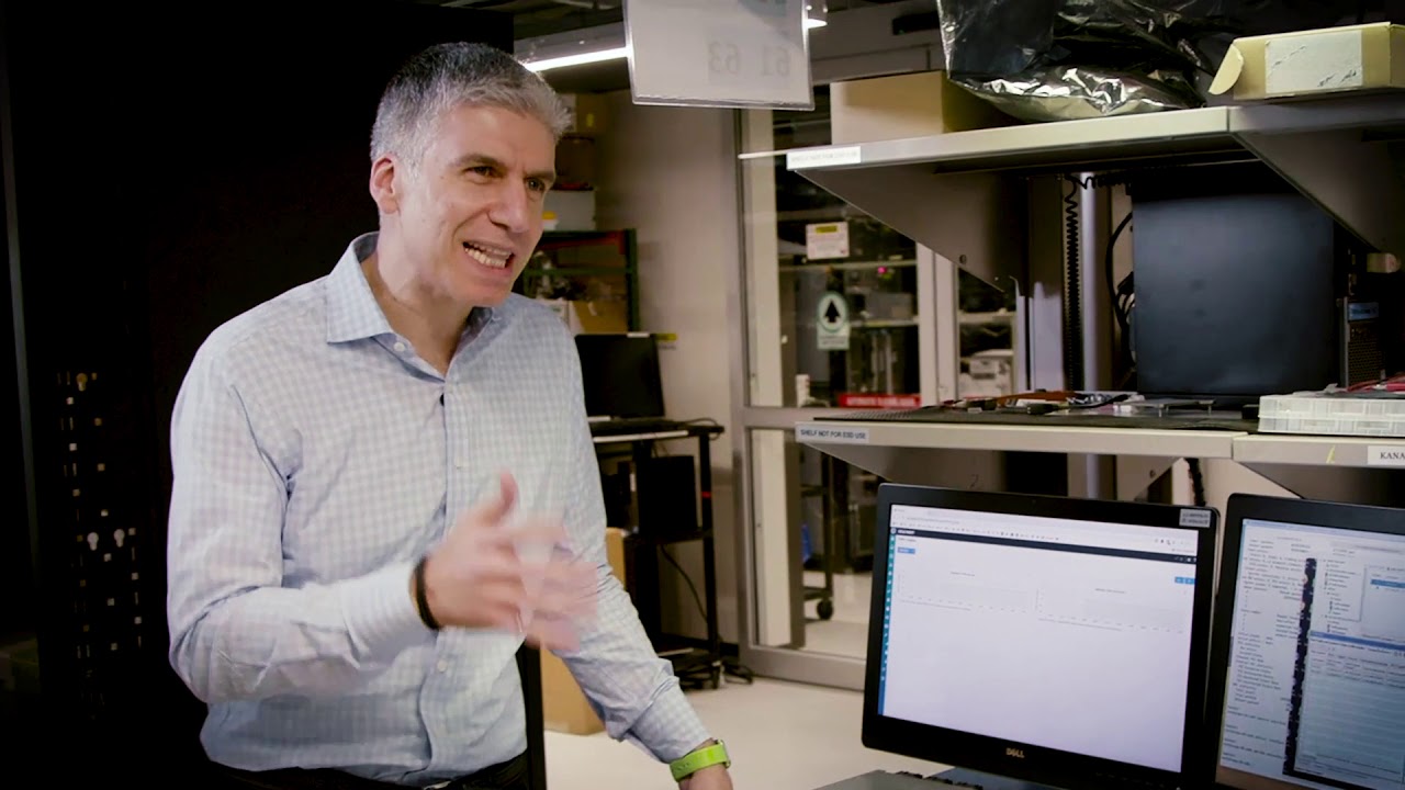 Rami Rahim, CEO, Juniper Networks, standing next to shelves with computer monitors and hardware parts, gesturing while talking in a video to someone offscreen about Juniper router capabilities.